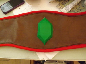 A photo of the center of a wide brown belt with red trim and a green rupee in the middle.