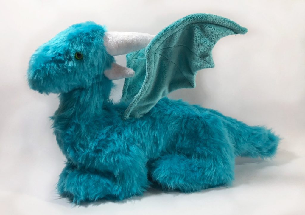 Photo of a teal, faux fur dragon plush with wired wings, as seen from the side. It is sitting down on four legs, with wings unfurled.