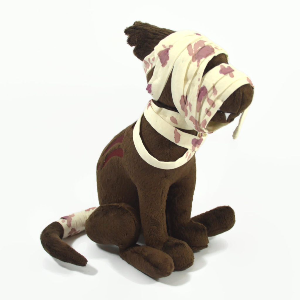 A brown zombie dog plush. It is sitting upright and has bloody bandages on its head, shoulders, and tail. It has red embroidered gashes in its side and jagged teeth are visible through the bandages on the muzzle.