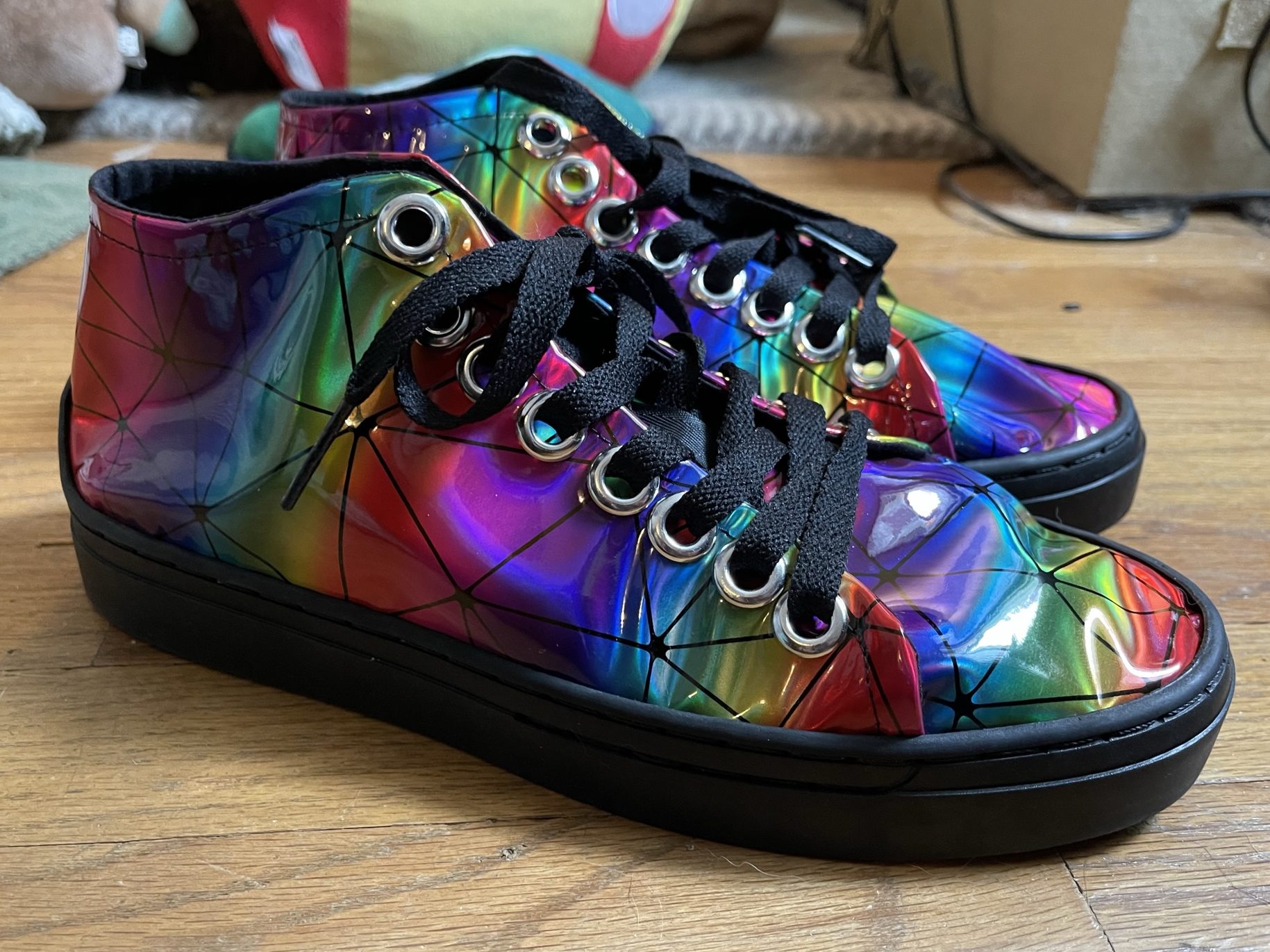 A photo of a pair of high-top sneakers made out of shiny rainbow vinyl with black rubber soles and black laces.