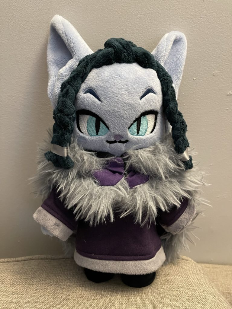 A photo of a chibi-style plush doll of Grabalba from Dimension 20. She is light blue with dark green hair and purple and gray dress and cloak. She has big bat ears and embroidered teal eyes.