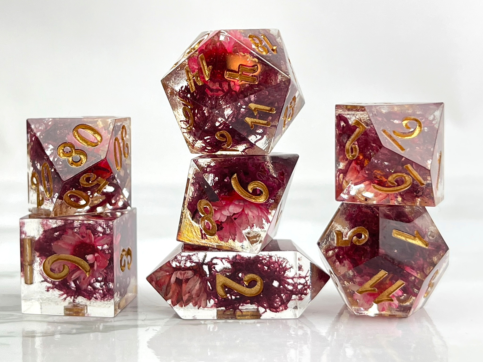 Photo of a set of seven dice that are clear resin with red moss and flowers inside. The numbers are inked in gold.