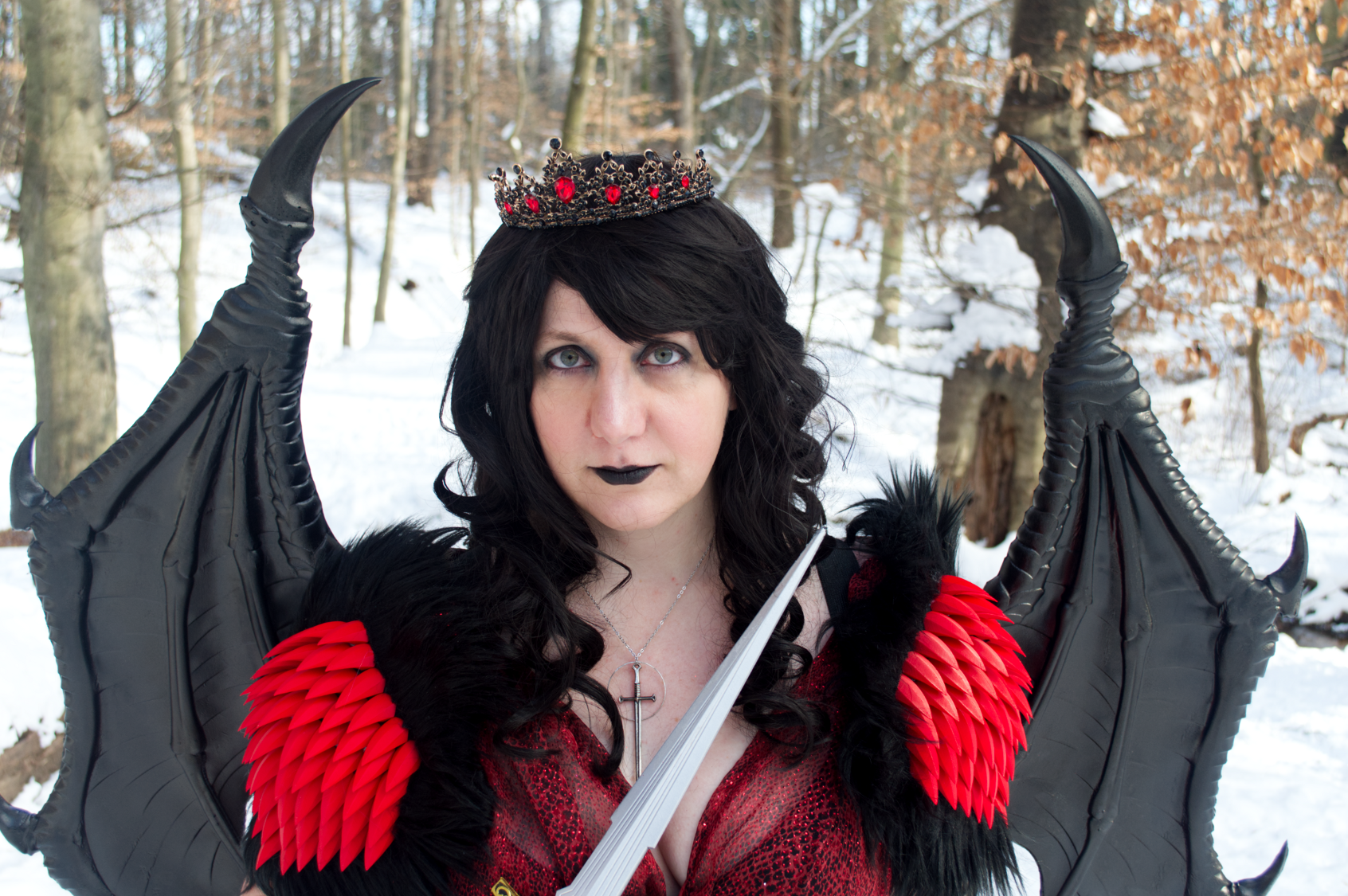 Cosplay portrait of a woman with long dark wavy hair wearing a crown, a red snakeprint dress, shoulder armor with spikes and fur, and dragon wings.