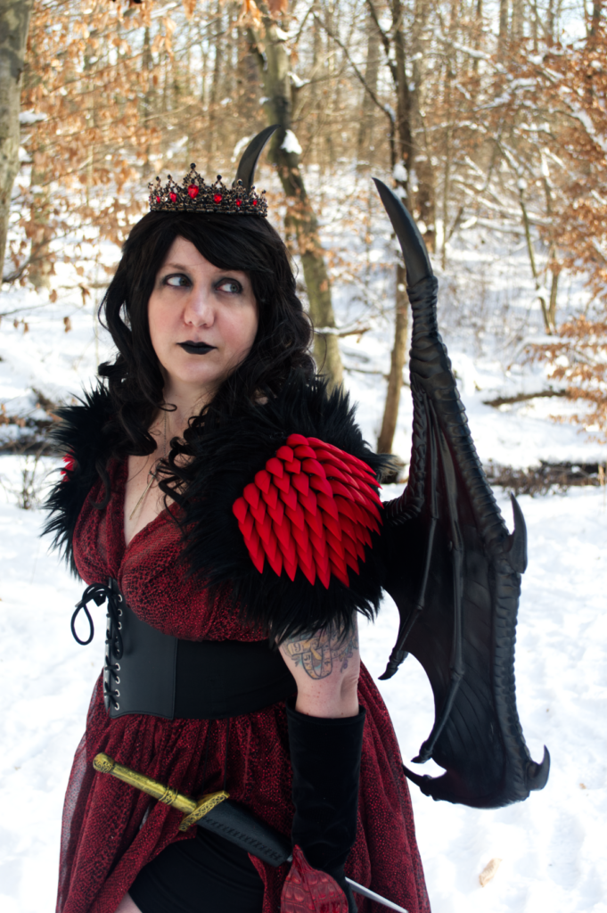 Cosplay portrait of a woman with long dark wavy hair wearing a crown, a red snakeprint dress, shoulder armor with spikes and fur, and dragon wings. She has a dagger in a frog at her waist.