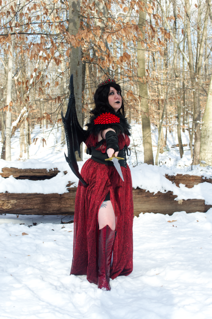Cosplay portrait of a woman with long dark wavy hair wearing a crown, a red snakeprint dress, shoulder armor with spikes and fur, and dragon wings. She is pointing a dagger at the viewer.