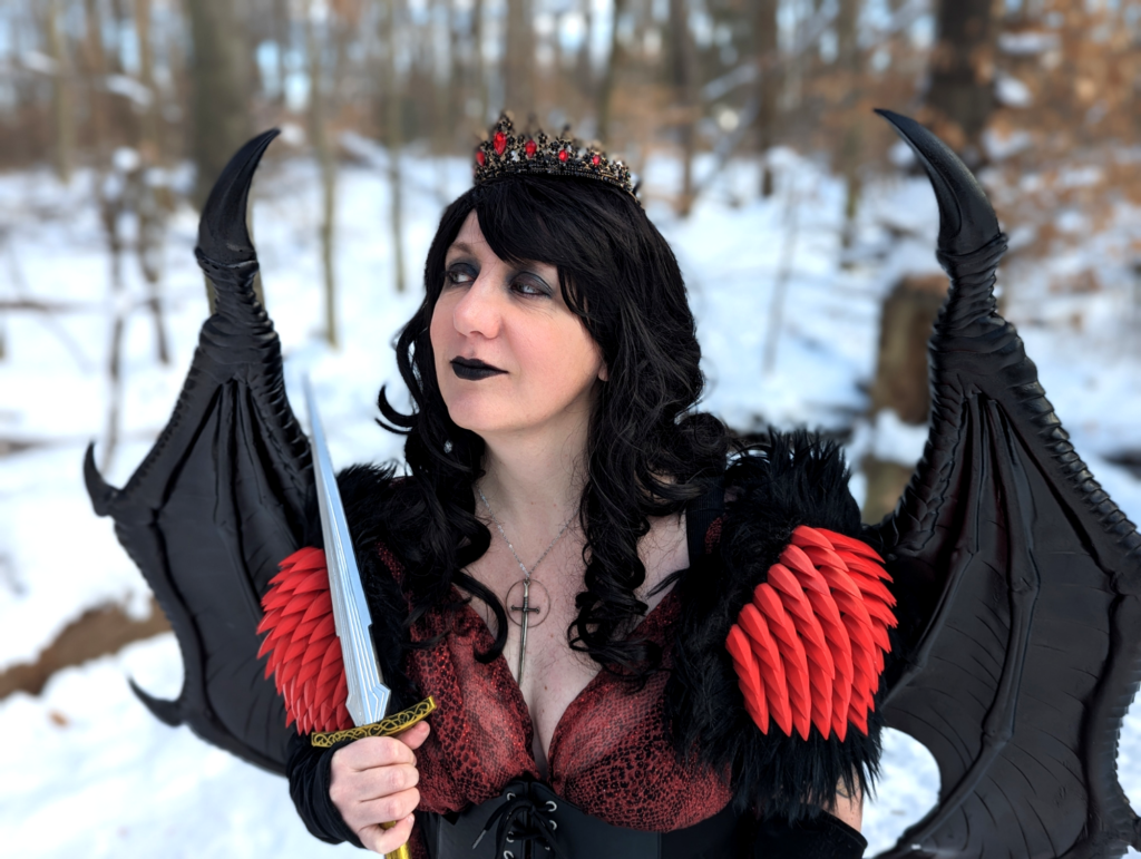 Cosplay portrait of a woman with long dark wavy hair wearing a crown, a red snakeprint dress, shoulder armor with spikes and fur, and dragon wings. She has a smug expression.