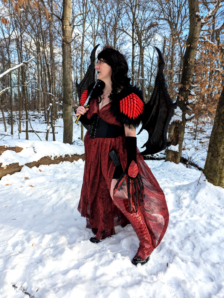 Cosplay portrait of a woman with long dark wavy hair wearing a crown, a red snakeprint dress, shoulder armor with spikes and fur, and dragon wings.
